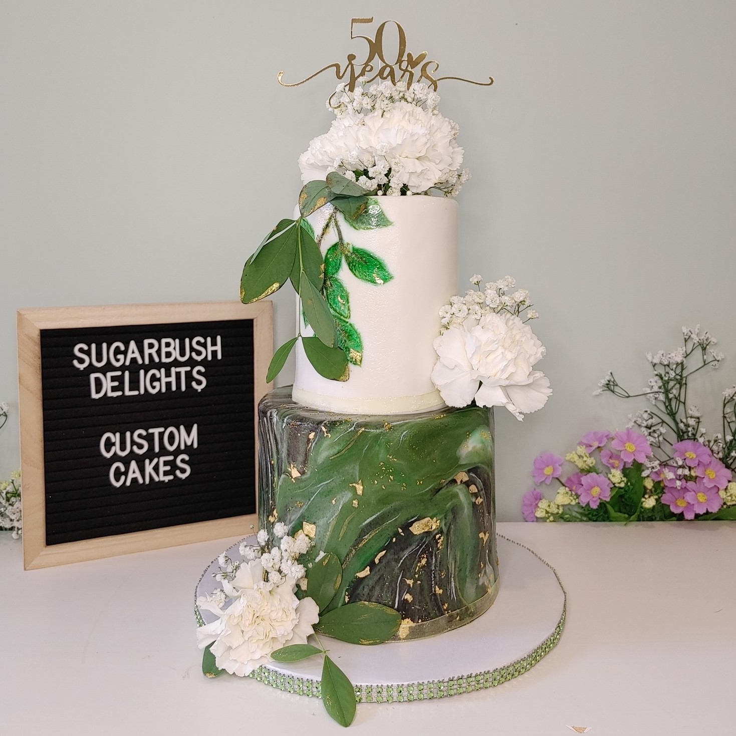 A tall 2 tier cake with white flowers and green leaves. The top tier is white and the bottom tier is green and black marble. There's a cake topper that says 50 years.