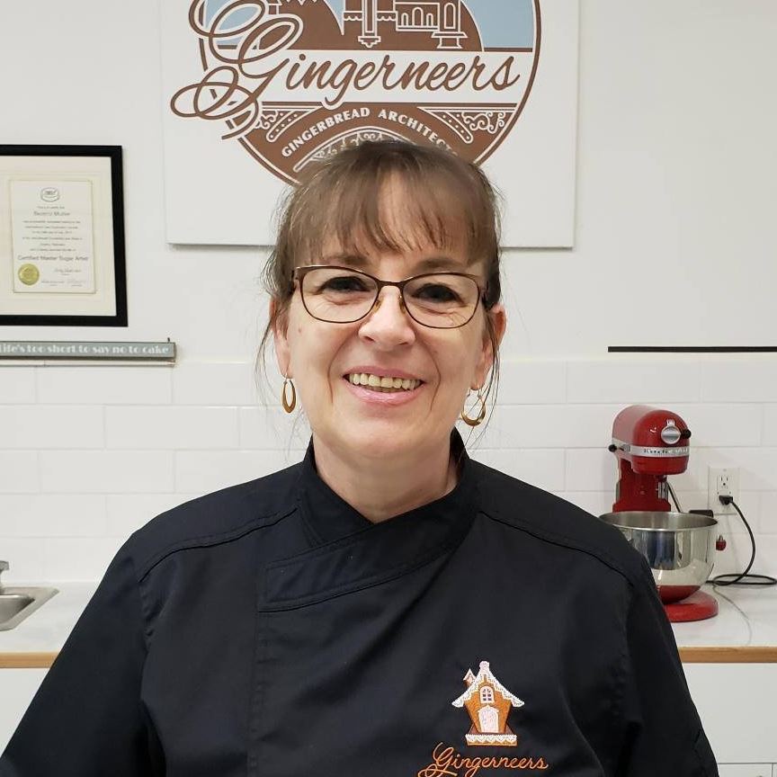 A picture of Beatriz Muller smiling in a black chefs jacket with the Gingerneers logo embroidered on it. She is standing in front of a white wall with a Gingerneers sign and sugar arts certificate hanging. There is a white counter with a red mixer on it to the right.
