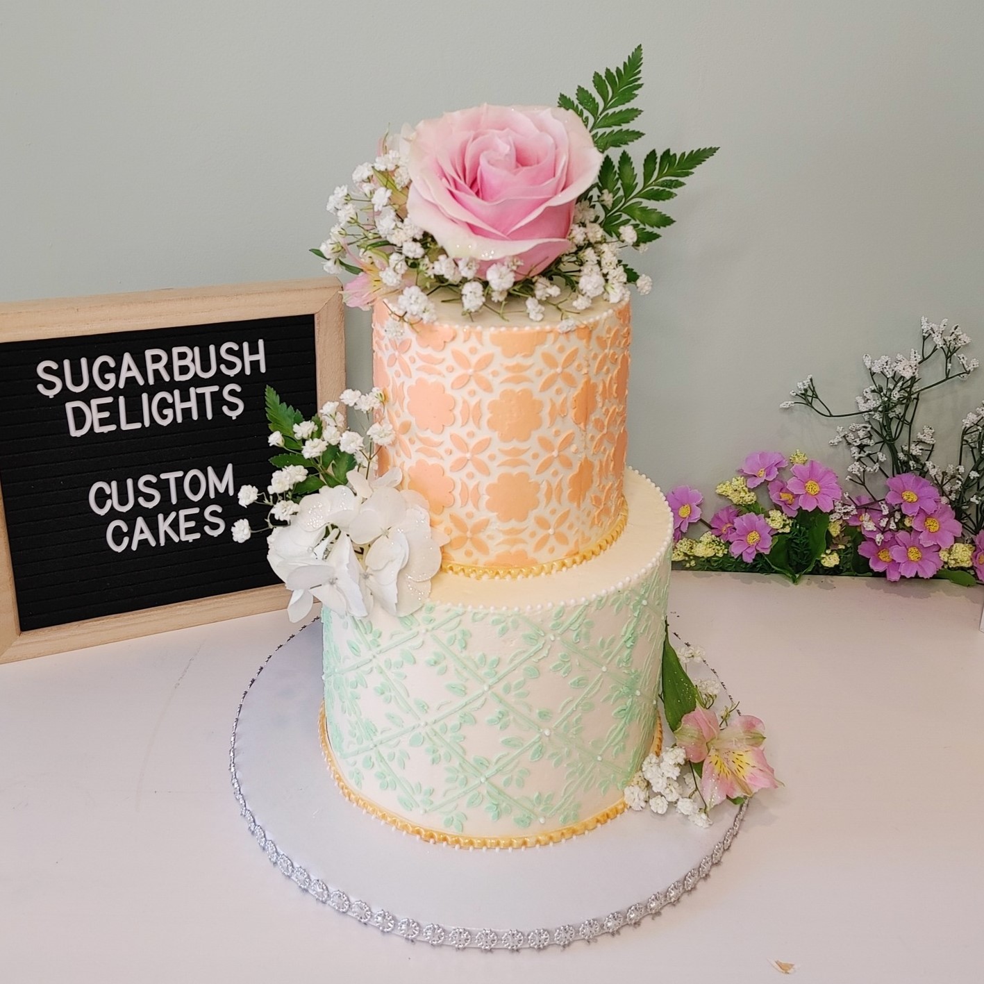 A 2 tier white cake with orange lace buttercream on the first tier and green on the second tier. There is a pink rose on top with white babys breath and 2 green leaves. There is a white flower with babys breath on top of the second tier.