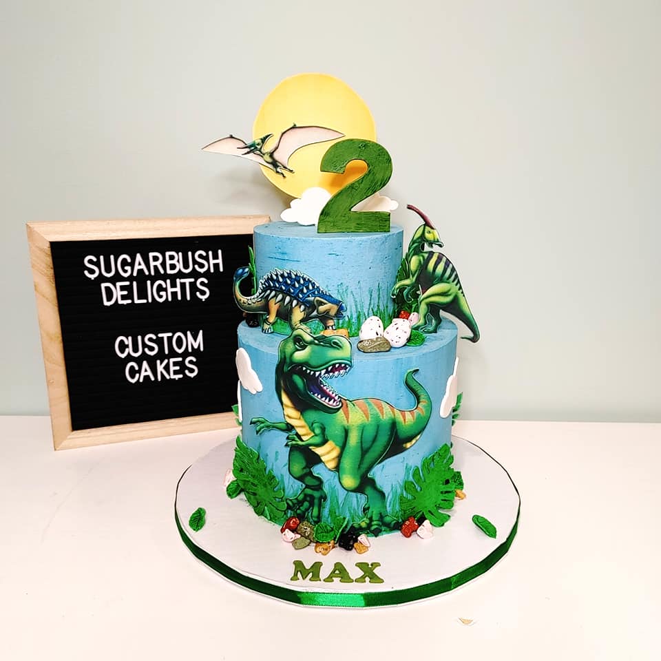 A 2 tier blue cake with dinosaurs and a number 2 on it. On the cake board it says Max.