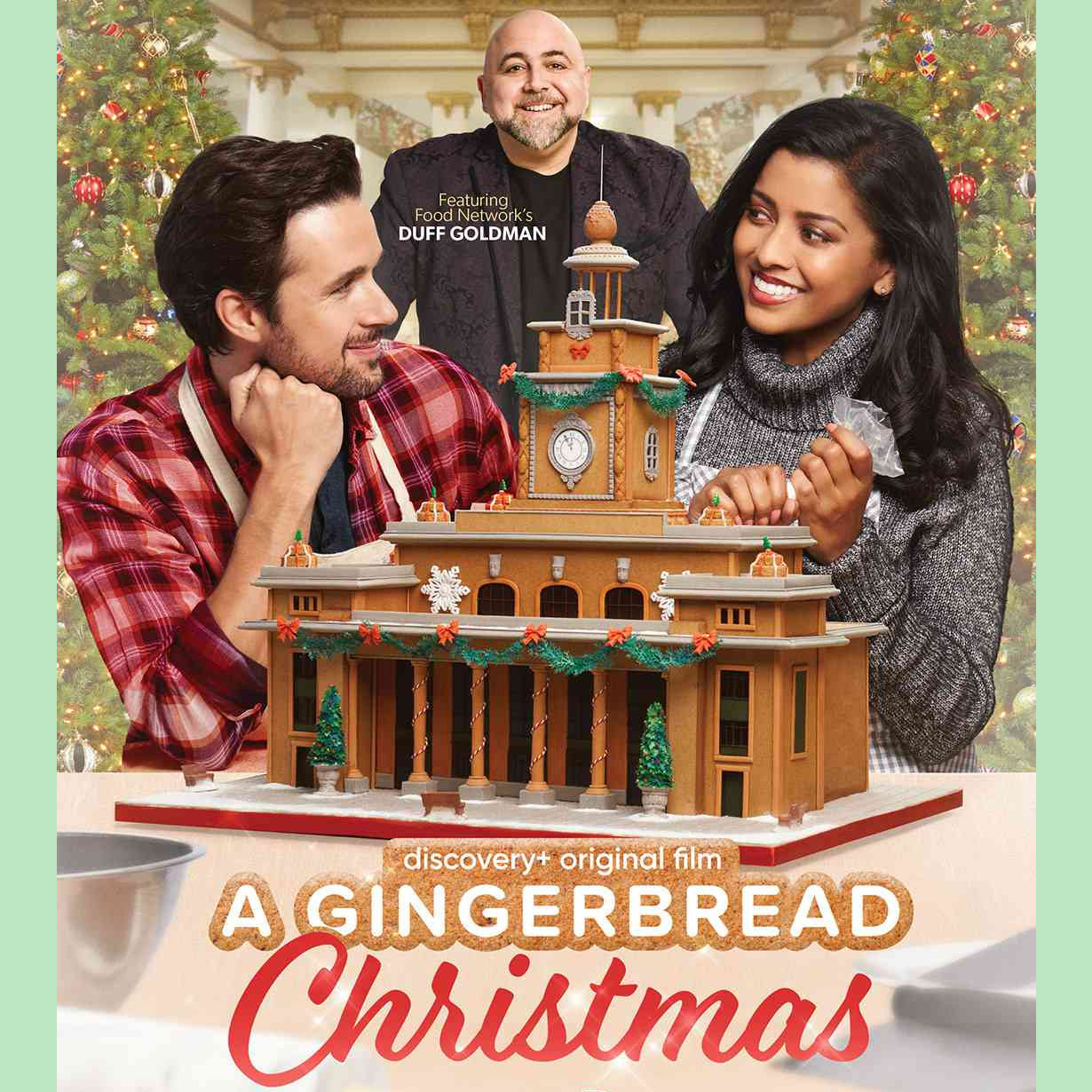 A Christmas movie poster. James is wearing a red flannel top and is staring lovingly at Hazel with his head resting on his chin. Hazel is wearing a grey sweater and a apron, holding a piping bag, smiling at James. They are sitting behind a beautiful gingerbread city hall. Duff Goldman is behind them. The title 'A Gingerbread Christmas' is at the bottom of the poster.