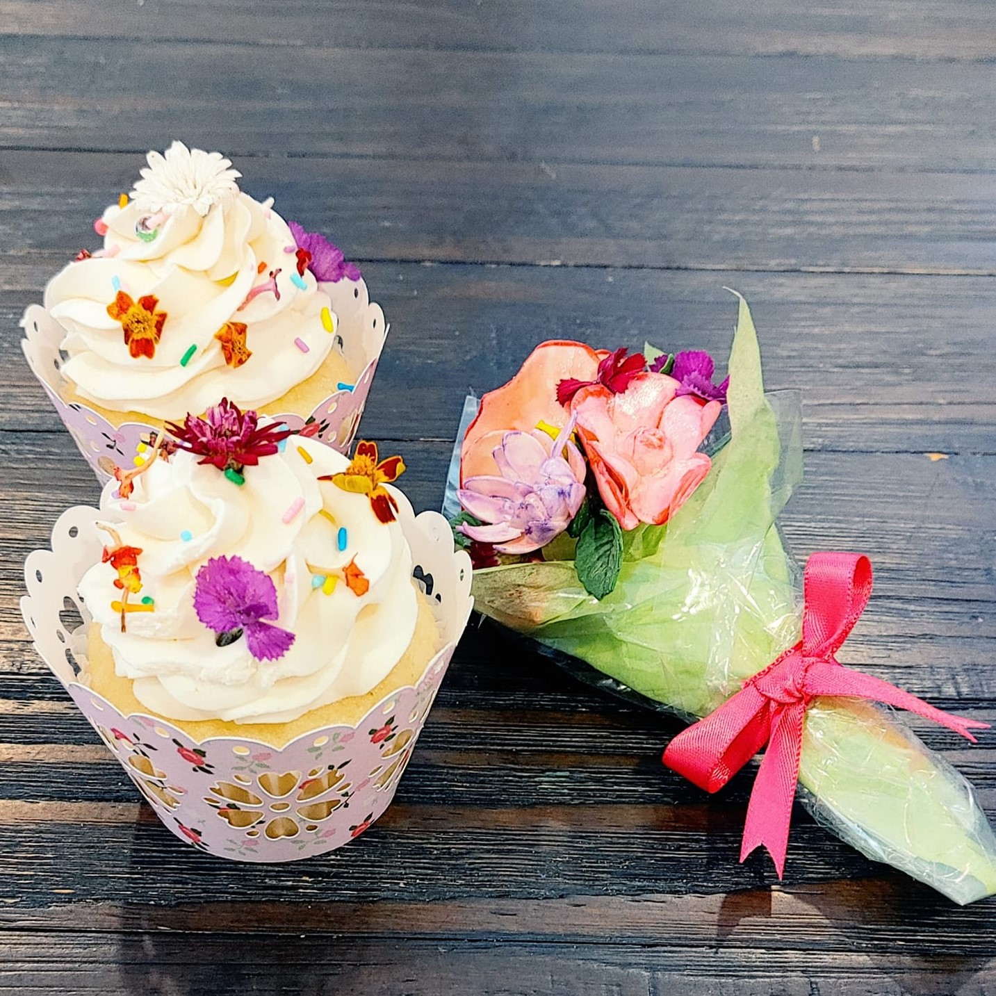 2 white cupcakes with tiny real flowers on them in a pink cupcake liner. There is a small bouquet of flowers beside it in a green wrapper and a pink bow.